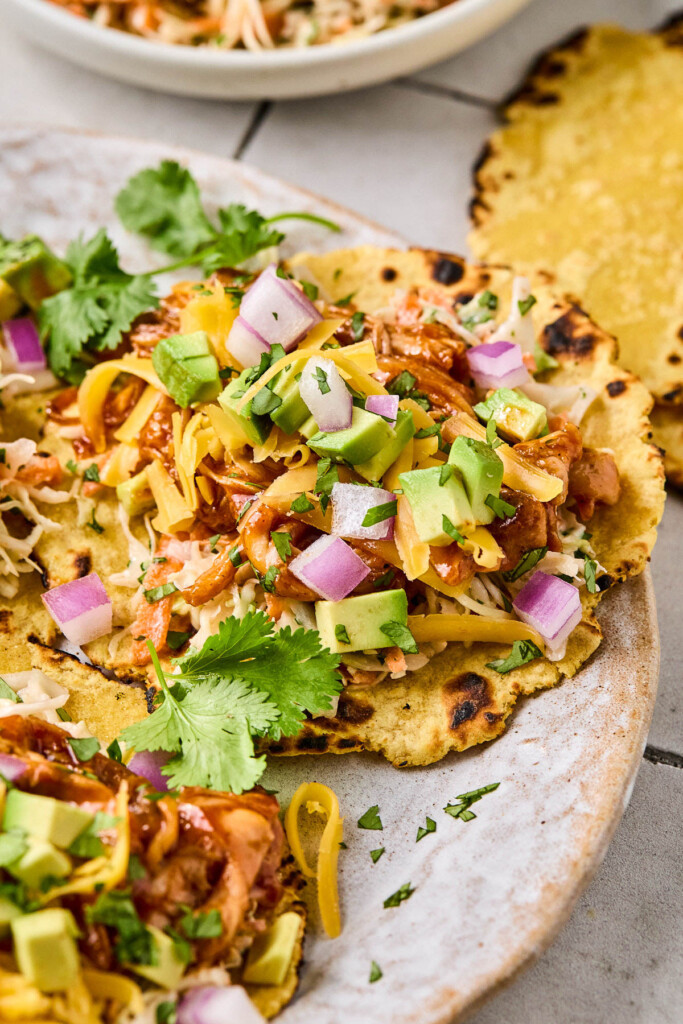 Barbecue chicken tacos on a plate with slaw, diced avocado, shredded cheddar cheese, diced red onion, and cilantro in a corn tortilla. Next to the plate is a corn tortilla.