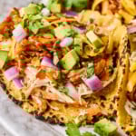 Bbq chicken tacos in a corn tortilla with coleslaw, shredded bbq chicken, avocado, shredded cheese, red onion, and cilantro on a plate.