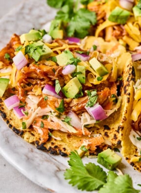 Bbq chicken tacos in a corn tortilla with coleslaw, shredded bbq chicken, avocado, shredded cheese, red onion, and cilantro on a plate.