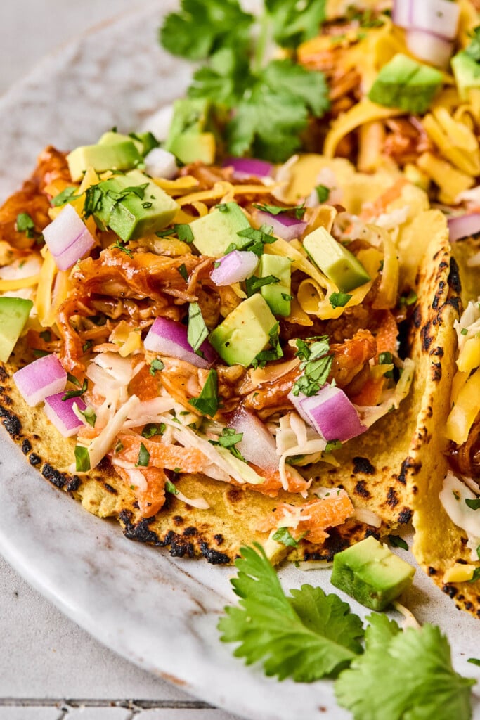 Bbq chicken tacos in a corn tortilla with coleslaw, shredded bbq chicken, avocado, shredded cheese, red onion, and cilantro on a plate.