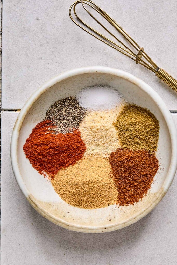 A small bowl filled with piles of different spices before they are mixed together. There is a small whisk next to the bowl.