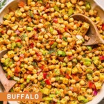 Buffalo chickpea salad with veggies, cheese, and herbs in a large bowl with two wooden spoons in it. Next to the bowl is fresh dill, half a lemon and juiced, and a jar of dressing.