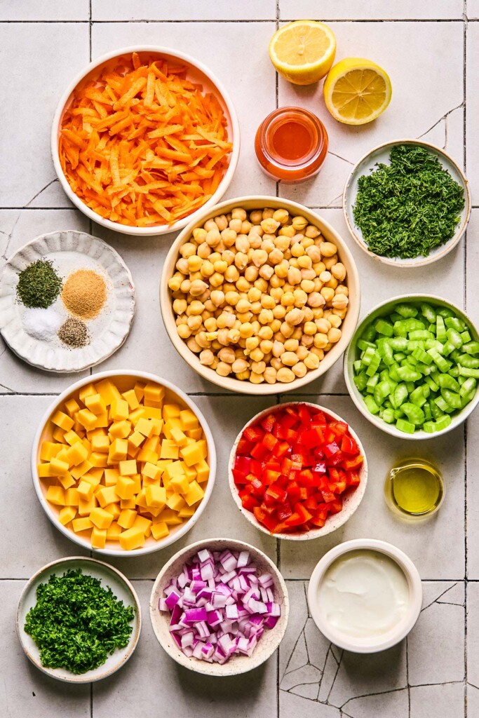Grey tile counter with a lemon that has been cut in half, a small jar of hot sauce, a small plate of chopped dill, a bowl of chickpeas, a bowl of diced celery, a bowl of diced red bell pepper, a small jar of olive oil, a bowl of greek yogurt, a bowl of diced red onion, a small plate of chopped parsley, a bowl of cubed cheddar cheese, a plate of a few piles of different spices, and a bowl of shredded carrots on it.