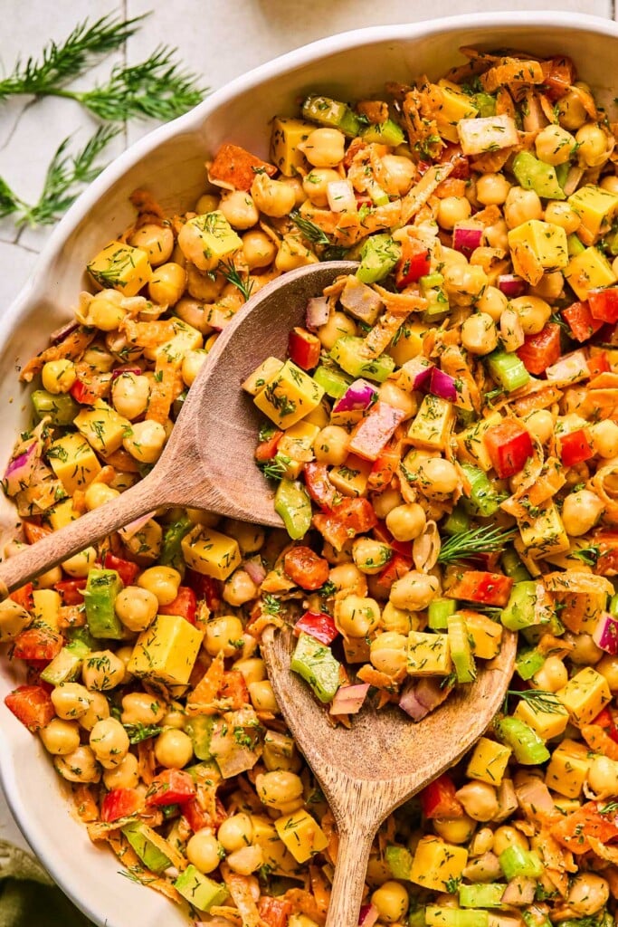 Healthy chickpea salad with bell peppers, carrots, celery, red onion, cheese, and herbs tossed in spicy dressing in a large bowl. There are 2 wooden spoons in the bowl and next to the bowl is fresh dill.