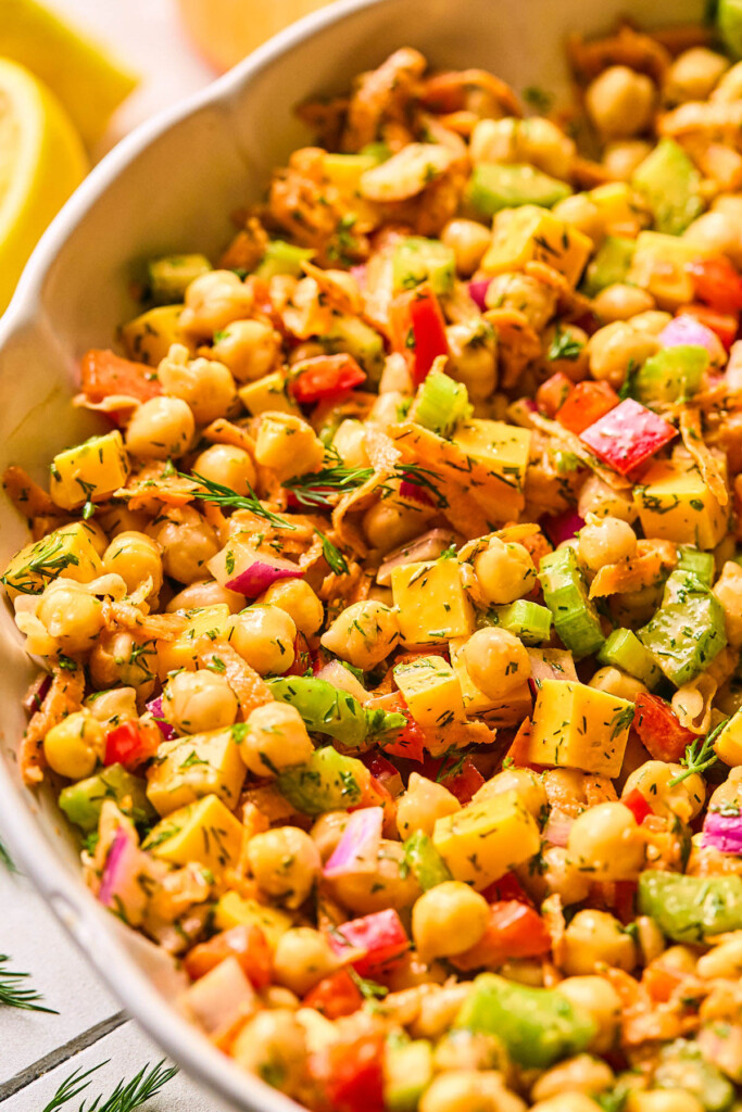 A close up of spicy chickpea salad with veggies, herbs, and cheese in a large white bowl. Off in the background are some lemons.
