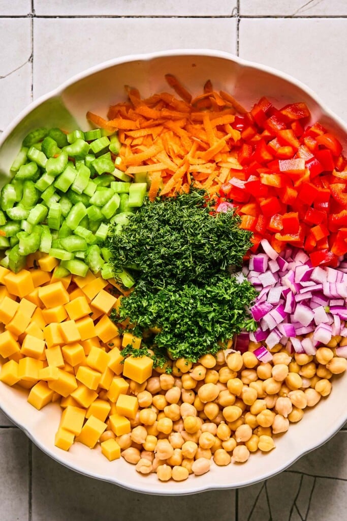 Large white bowl with chickpeas, cubed cheddar cheese, diced celery, shredded carrots, diced red bell pepper, diced red onion, and chopped fresh herbs before it is mixed together.