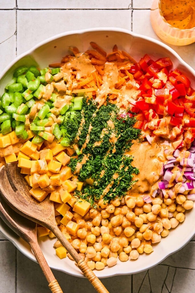 Large white bowl with chickpeas, cubed cheddar cheese, diced celery, shredded carrots, diced red bell pepper, diced red onion, and chopped fresh herbs with dressing poured over it before it is mixed together. There are wooden salad tongs in the bowl and next to the bowl is a jar of more dressing.