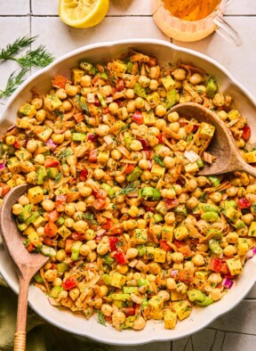 Buffalo chickpea salad with veggies, cheese, and herbs in a large bowl with two wooden spoons in it. Next to the bowl is fresh dill, a lemon that has been cut in half and juiced, a jar of dressing, and a green kitchen linen.