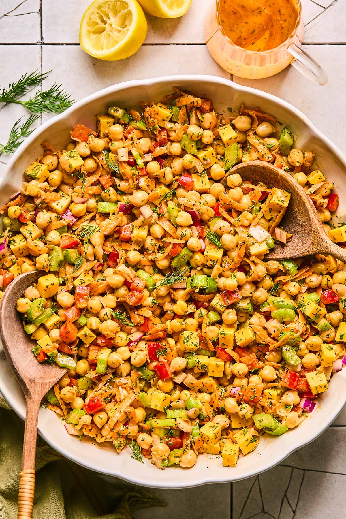 Buffalo chickpea salad with veggies, cheese, and herbs in a large bowl with two wooden spoons in it. Next to the bowl is fresh dill, a lemon that has been cut in half and juiced, a jar of dressing, and a green kitchen linen.