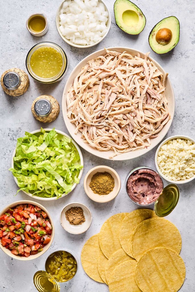 Grey concrete counter with an avocado cut in half, a large bowl of shredded chicken, a bowl of crumbled queso fresco, an opened can of refried black beans, corn tortillas, two small bowls of spices, an opened can of diced green chiles, a bowl of pico de gallo, a bowl of shredded lettuce, a salt and pepper shaker, a bowl of green salsa, and a small container of olive oil.