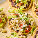 Chicken tostadas with refried beans, seasoned shredded chicken, lettuce, pico de gallo, queso fresco, and cilantro on them. They are on a baking sheet lined with parchment paper.