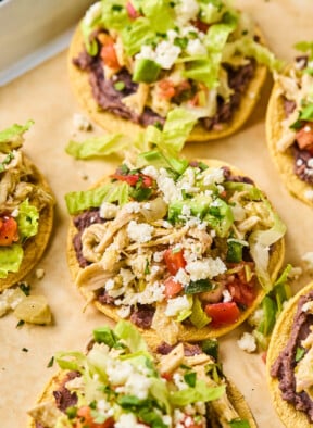 Chicken tostadas with refried beans, seasoned shredded chicken, lettuce, pico de gallo, queso fresco, and cilantro on them. They are on a baking sheet lined with parchment paper.