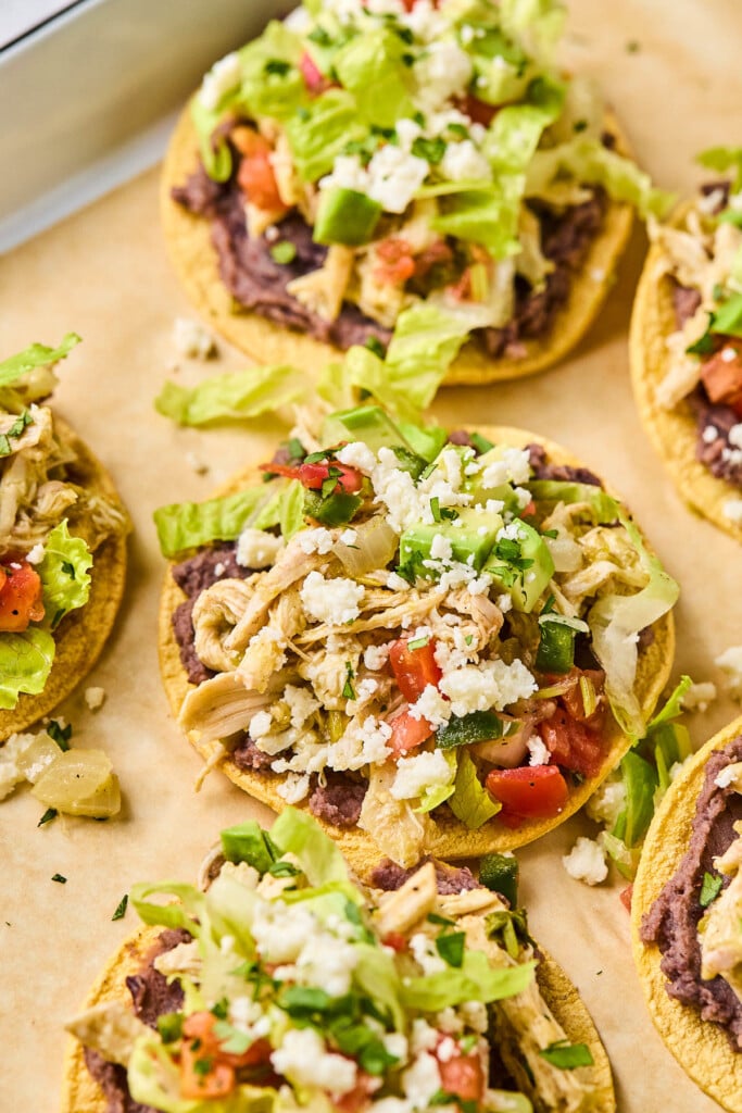 Chicken tostadas with refried beans, seasoned shredded chicken, lettuce, pico de gallo, queso fresco, and cilantro on them. They are on a baking sheet lined with parchment paper.