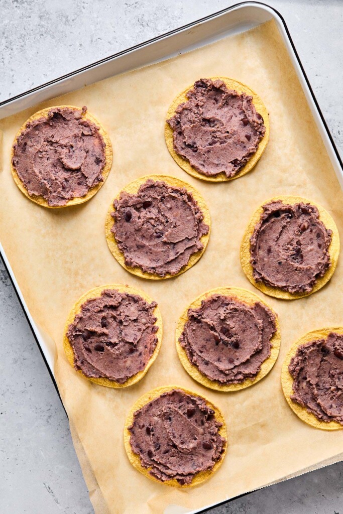 Overhead shot of a baking sheet lined with parchment paper with crispy corn tortilla shells with refried beans spread on them.