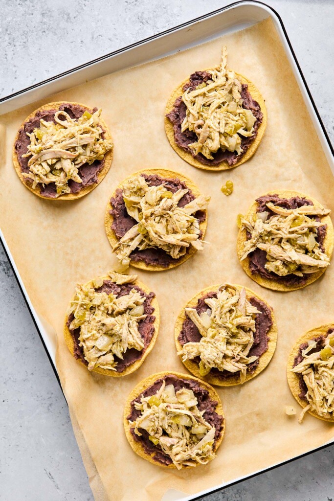 Overhead shot of a baking sheet lined with parchment paper with crispy corn tortilla shells with refried beans and seasoned shredded chicken on them.