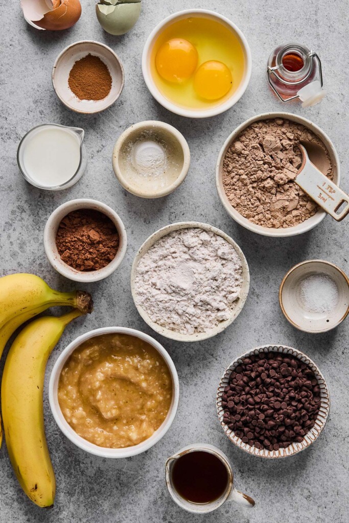 Grey concrete counter with a bottle of vanilla extract, a bowl of chocolate protein powder with a measuring spoon in it, a small dish of salt, a bowl of mini chocolate chips, a small cup of maple syrup, a bowl of mashed bananas, two ripe bananas, a bowl of cocoa powder, a small bowl of baking powder, a cup of milk, a small bowl of cinnamon, two eggs that have been cracked into a bowl, and two egg shells.