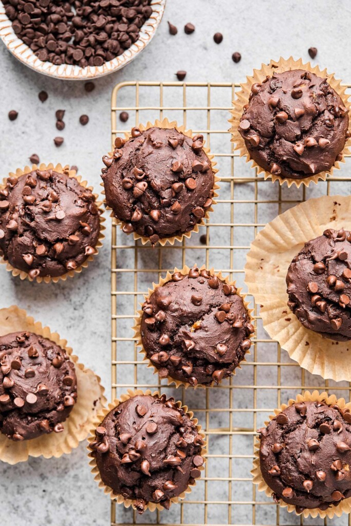 Overhead shot of chocolate protein muffins on a cooling rack. Next to the rack is a bowl of mini chocolate chips with some of them spilled next to the bowl.