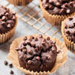 Chocolate protein muffin with some mini chocolate chips around it. Behind it is a cooling rack of more muffins.