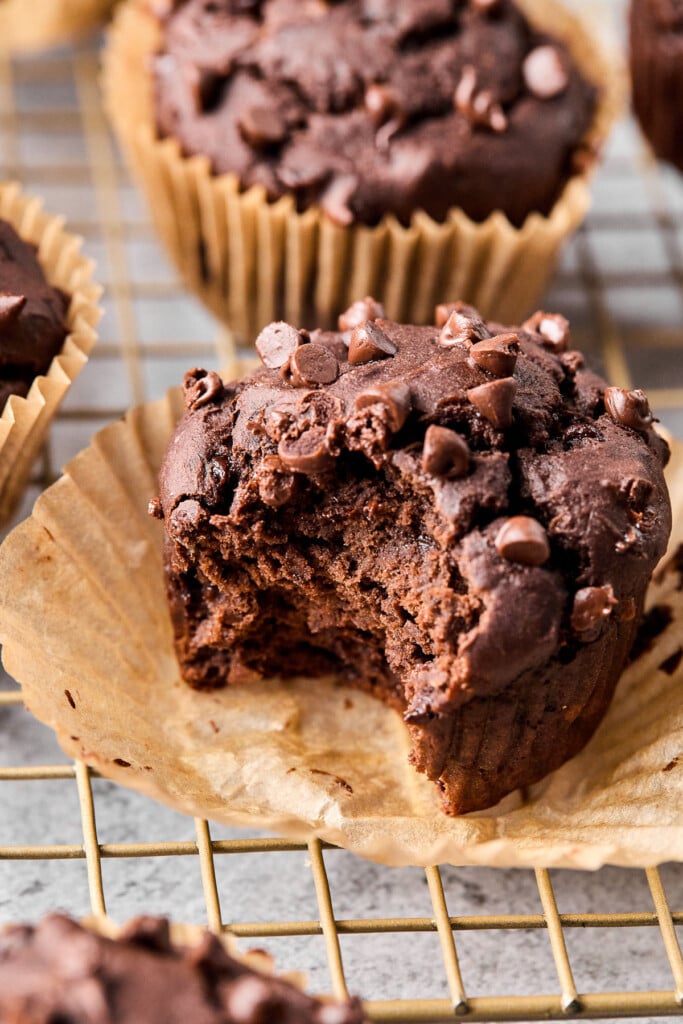 Chocolate banana protein muffin on a cooling rack with a bite taken out of it. There are more muffins behind it.