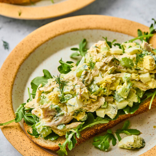Chicken and egg salad on top of some arugula on a piece of toast on a plate. It is topped with fresh dill. In front of the plate is a striped kitchen towel and behind the plate is another plate with chicken salad on toast on it.