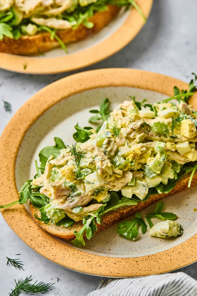 Chicken and egg salad on top of some arugula on a piece of toast on a plate. It is topped with fresh dill. In front of the plate is a striped kitchen towel and behind the plate is another plate with chicken salad on toast on it.