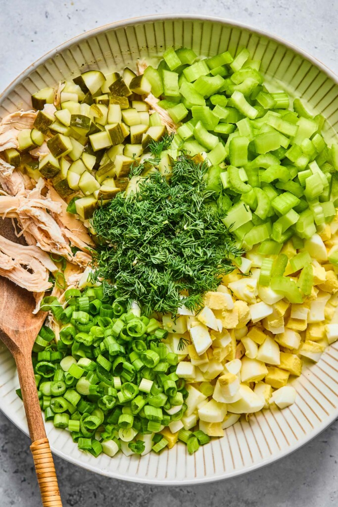 Large bowl with diced celery, chopped hard-boiled eggs, sliced green onions, shredded chicken, diced pickles, and fresh dill in separate piles before they are mixed together. A wooden spoon is in the bowl.
