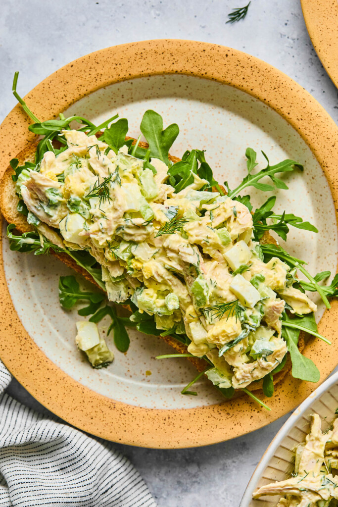 Chicken salad recipe with eggs and celery on top of some arugula on a piece of toast on a plate. Next to the plate is a striped kitchen towel.
