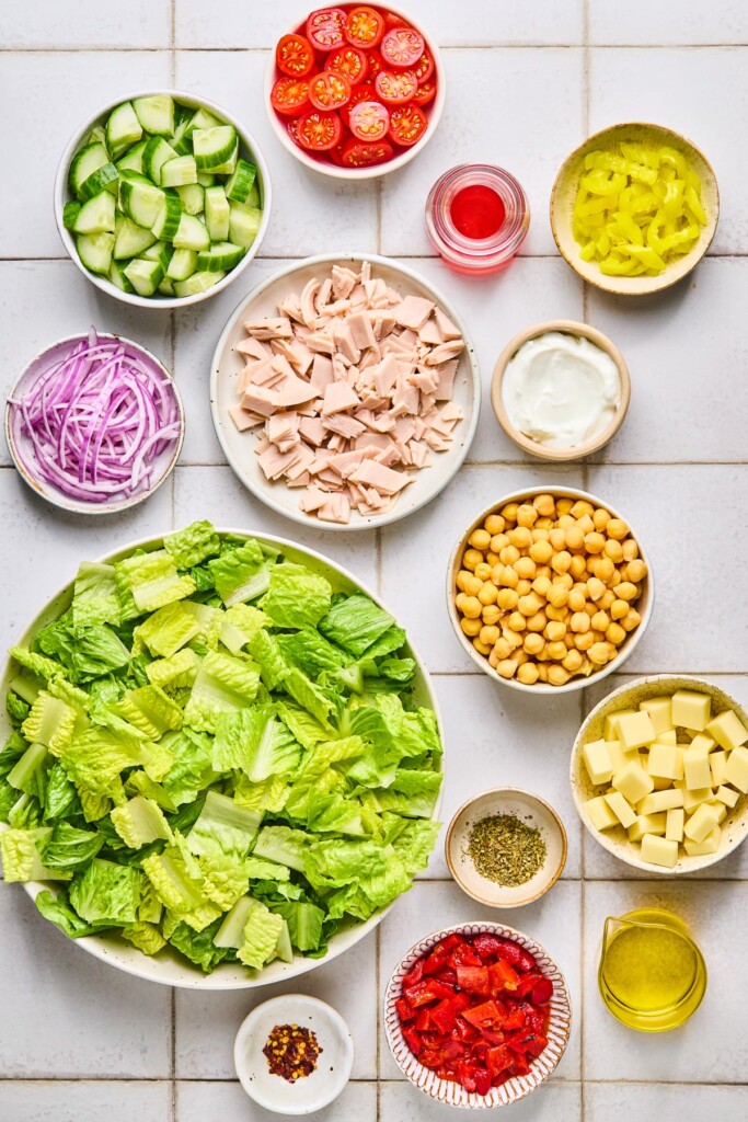 White tile counter with a bowl of halved cherry tomatoes, a small jar of red wine vinegar, a small bowl of banana peppers, a bowl of chopped deli turkey, a small bowl of greek yogurt, a bowl of chickpeas, a bowl of cubed cheese, a small dish of spices, a jar of olive oil, a small bowl of diced roasted red peppers, a small dish of red pepper flakes, a bowl of chopped romaine, a bowl of sliced red onion, and a bowl of diced cucumbers on it.