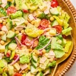 Chopped salad with lettuce, tomatoes, cucumber, turkey, cheese, chickpeas, red onion, and banana peppers in a large bowl. It is garnished with pepper and there is a large gold spoon in the bowl as well.