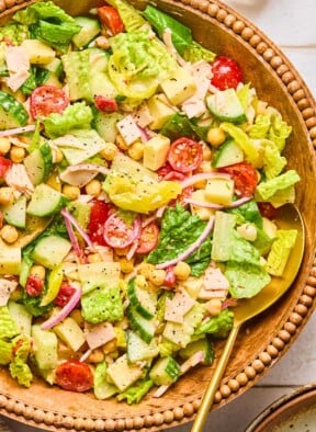 Chopped salad with lettuce, tomatoes, cucumber, turkey, cheese, chickpeas, red onion, and banana peppers in a large bowl. It is garnished with pepper and there is a large gold spoon in the bowl as well.
