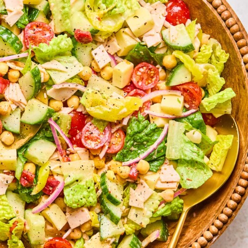 Chopped salad with lettuce, tomatoes, cucumber, turkey, cheese, chickpeas, red onion, and banana peppers in a large bowl. It is garnished with pepper and there is a large gold spoon in the bowl as well.