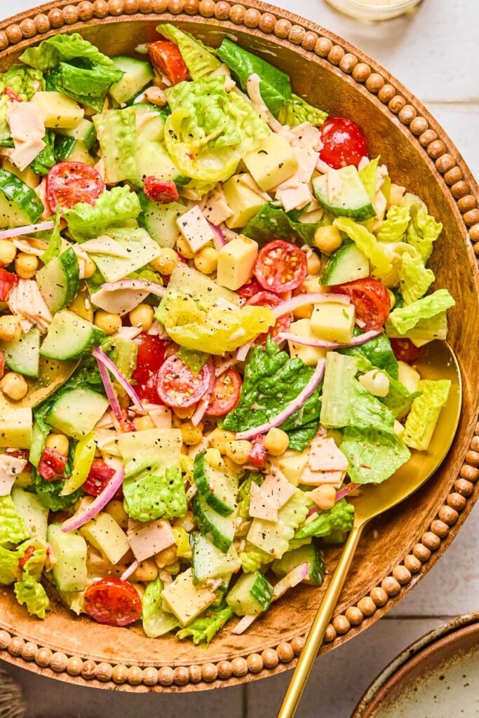 Chopped salad with lettuce, tomatoes, cucumber, turkey, cheese, chickpeas, red onion, and banana peppers in a large bowl. It is garnished with pepper and there is a large gold spoon in the bowl as well.