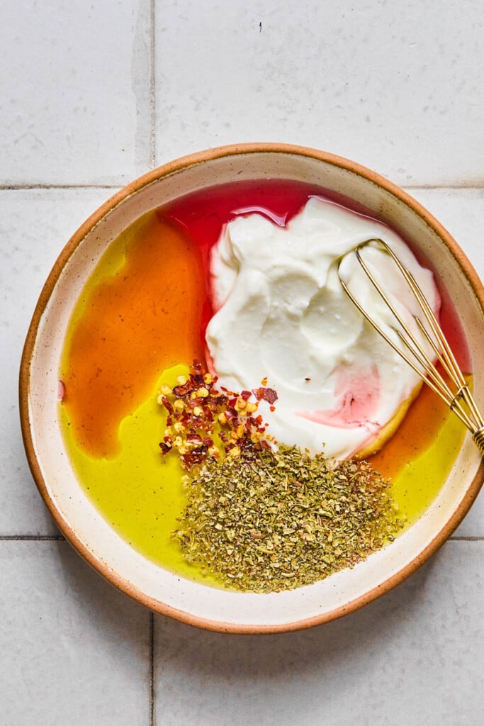 Small bowl with greek yogurt, red wine vinegar, olive oil, Italian seasoning, and red pepper flakes before it is mixed together. A small whisk is in the bowl as well.