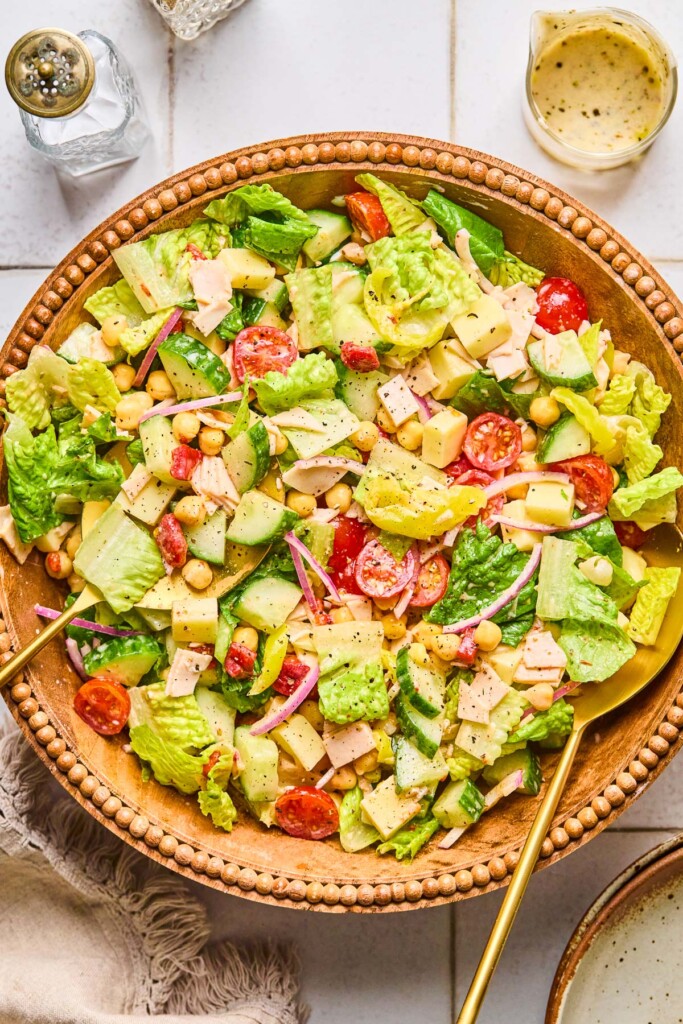Overhead shot of a large brown bowl with a chopped salad in it. The salad has lettuce, tomatoes, cucumber, turkey, cheese, chickpeas, red onion, and banana peppers in it. There is a large gold spoon in the bowl and next to the bowl is a tan linen kitchen towel, a salt and pepper shaker, and a jar with dressing in it.