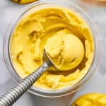 Ninja Creami pint container with mango frozen greek yogurt in it. A filled ice cream scoop is in the container. Around the container is fresh mango sliced from the seed.