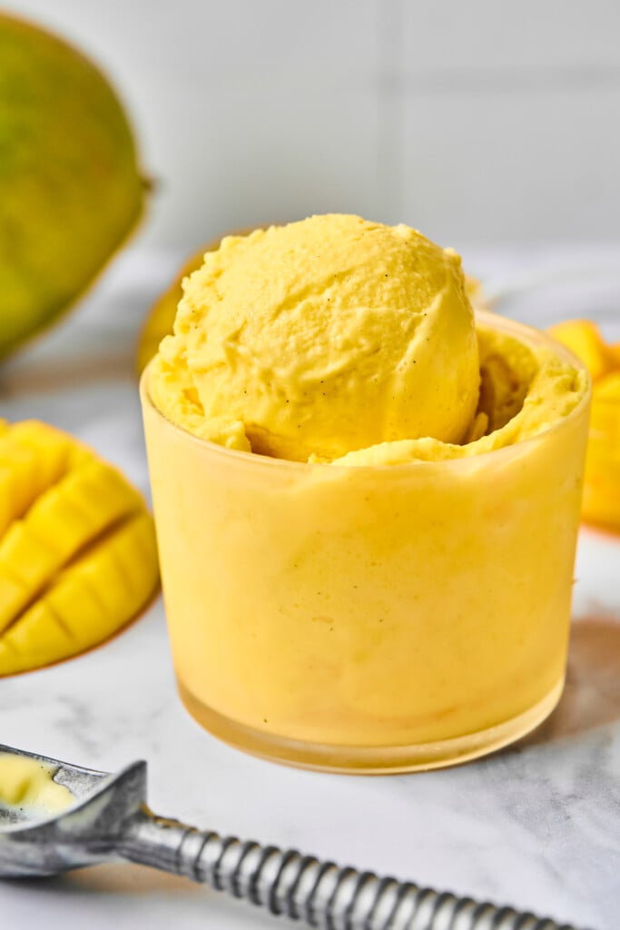 Scoops of mango ninja creami greek yogurt in a glass cup. Around the cup is an ice cream scoop, fresh mango cut off the seed, and a whole mango.
