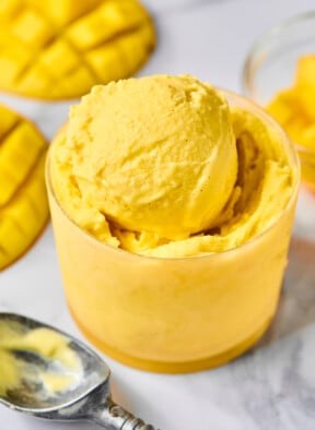 Scoops of mango Ninja Creami frozen yogurt in a small glass cup. Around the cup is an ice cream scoop and fresh mango that has been cut from the seed.