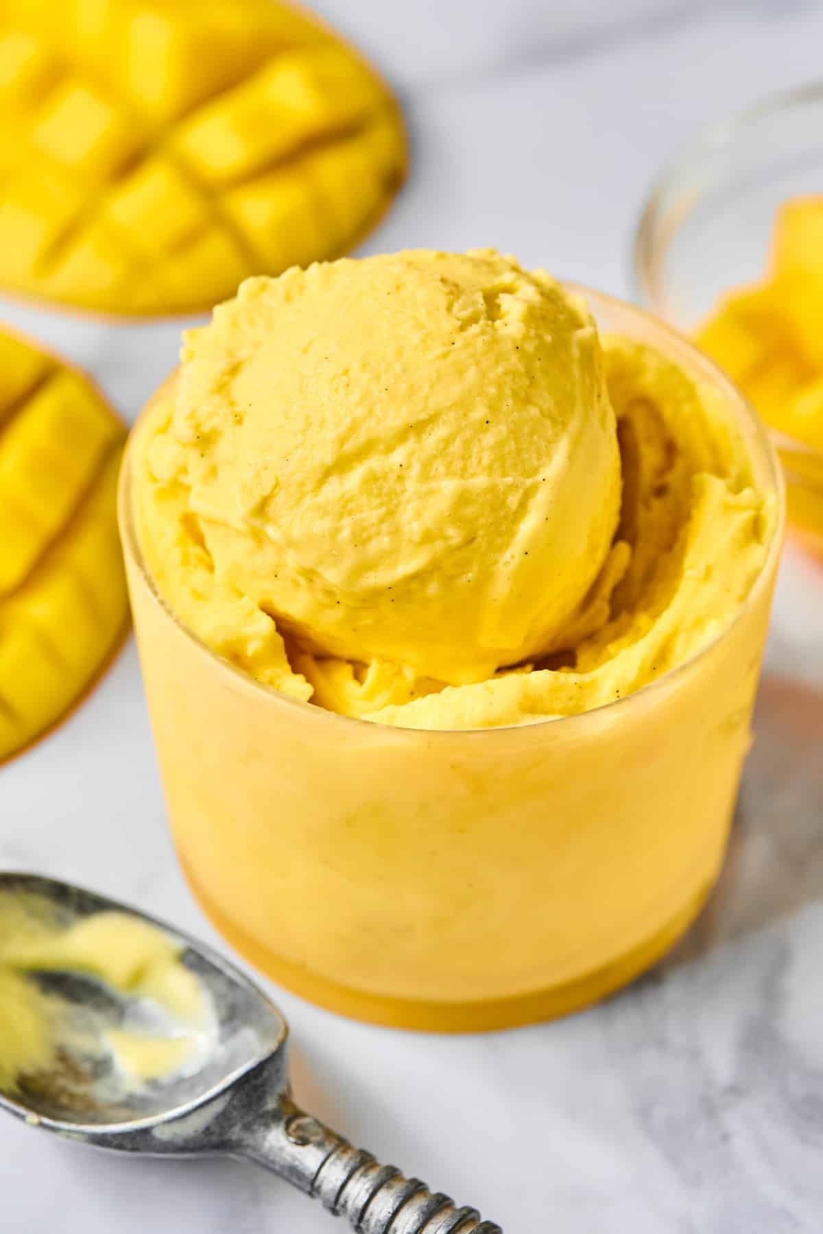 Scoops of mango Ninja Creami frozen yogurt in a small glass cup. Around the cup is an ice cream scoop and fresh mango that has been cut from the seed.