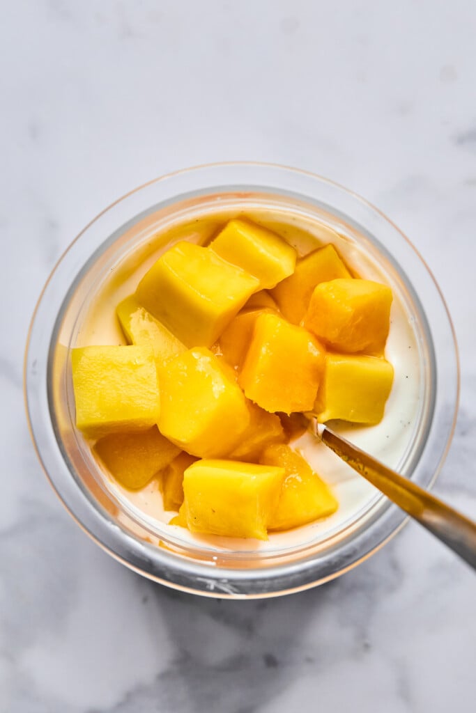 Diced mango overtop a greek yogurt mixture in a pint container. There is a spoon in the container as well.