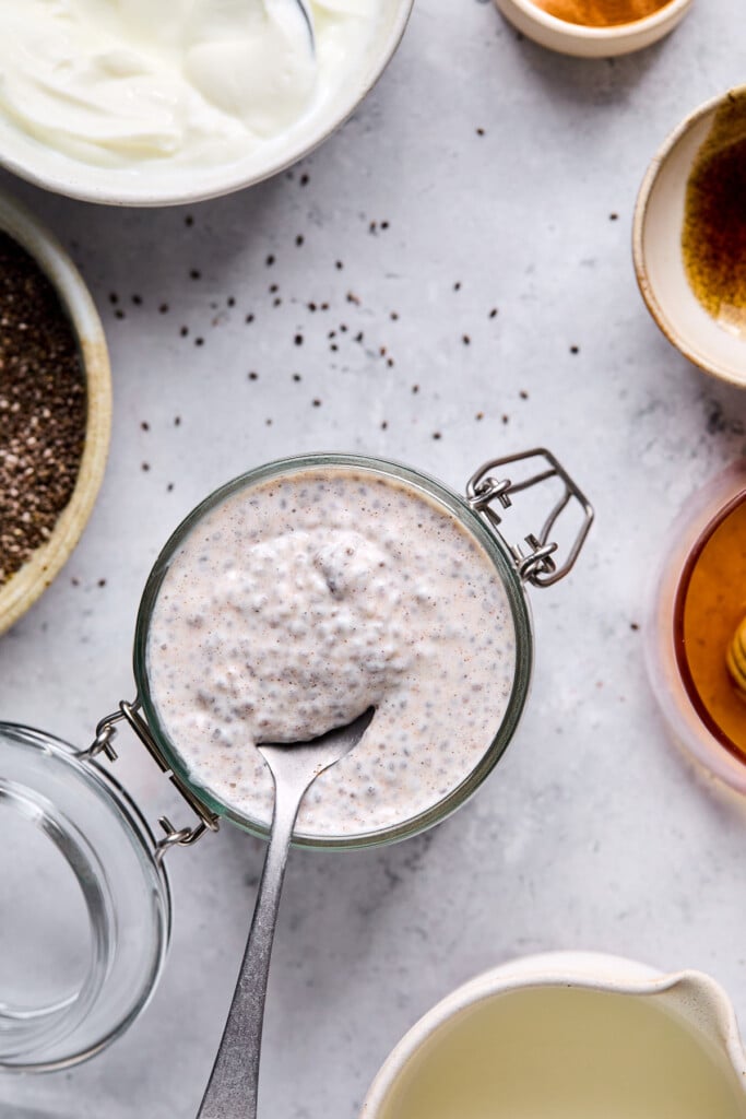 Greek yogurt chia pudding in a jar with a spoon in it. Around the jar is a bowl of greek yogurt, a bowl of vanilla bean paste, a bowl of honey, a cup of milk, and a bowl of chia seeds.