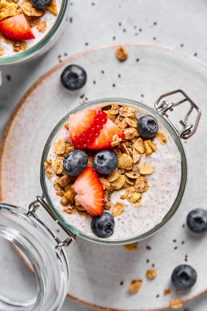 Overhead shot of protein chia seed pudding in a jar topped with strawberries, blueberries, and granola. The jar is sitting on a plate with a scattering of berries, chia seeds, and granola on it.