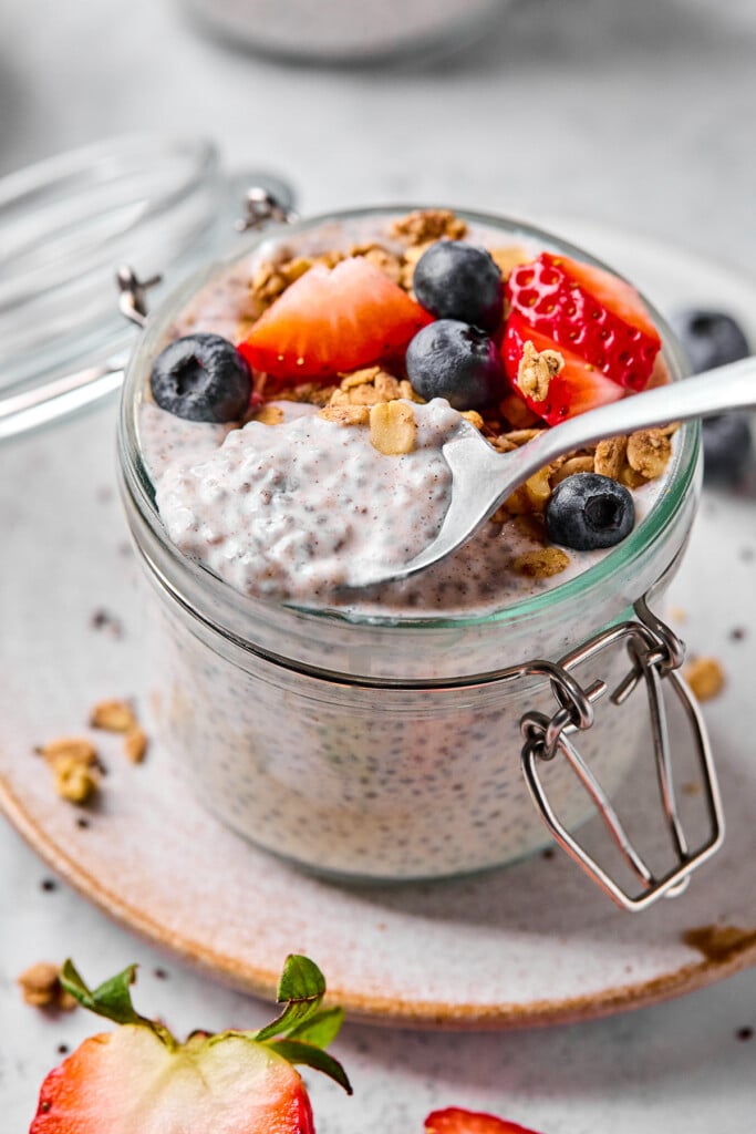 A jar of chia seed pudding with yogurt topped with strawberries, blueberries, and granola. A spoon is being scooped out of the jar and has chia pudding on it.