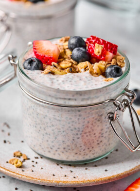 A jar of protein chia pudding topped with strawberries, blueberries, and granola. The jar is sitting on a plate and there is another jar filled with chia pudding behind the jar.