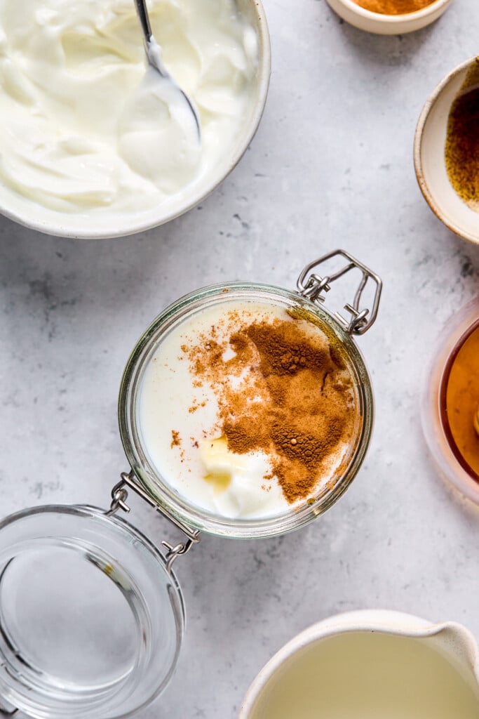 Overhead shot of a jar with greek yogurt, milk, honey, and cinnamon in it before it is mixed together. Around the jar is a bowl of greek yogurt, a bowl of vanilla bean paste, a bowl of honey, and a cup of milk.
