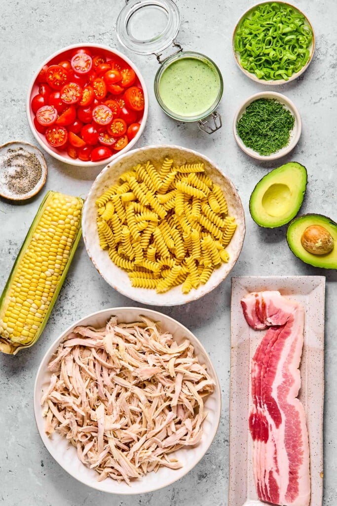 Grey concrete counter with a bowl of sliced green onions, a jar of jalapeño ranch, a small bowl of fresh dill, an avocado that has been cut in half, 4 pieces of uncooked bacon stacked on top of each other, a bowl of shredded chicken, a bowl of uncooked fusilli pasta, an ear of corn, a small bowl of salt and pepper, and a bowl of halved cherry tomatoes on it.