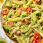 Chicken bacon ranch pasta salad in a large white bowl. There is a spoon in the bowl and it pasta salad is garnished with dill. Around the bowl is a small cup of jalapeño ranch and lemon slices.