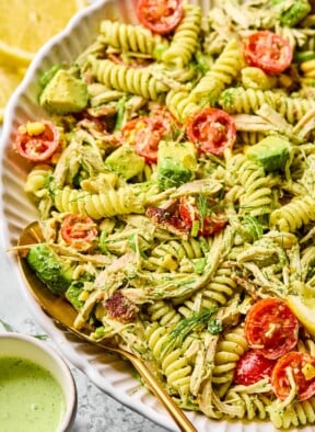 Chicken bacon ranch pasta salad in a large white bowl. There is a spoon in the bowl and it pasta salad is garnished with dill. Around the bowl is a small cup of jalapeño ranch and lemon slices.