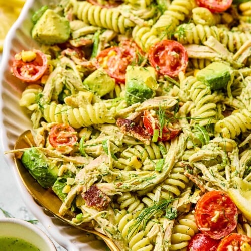 Chicken bacon ranch pasta salad in a large white bowl. There is a spoon in the bowl and it pasta salad is garnished with dill. Around the bowl is a small cup of jalapeño ranch and lemon slices.