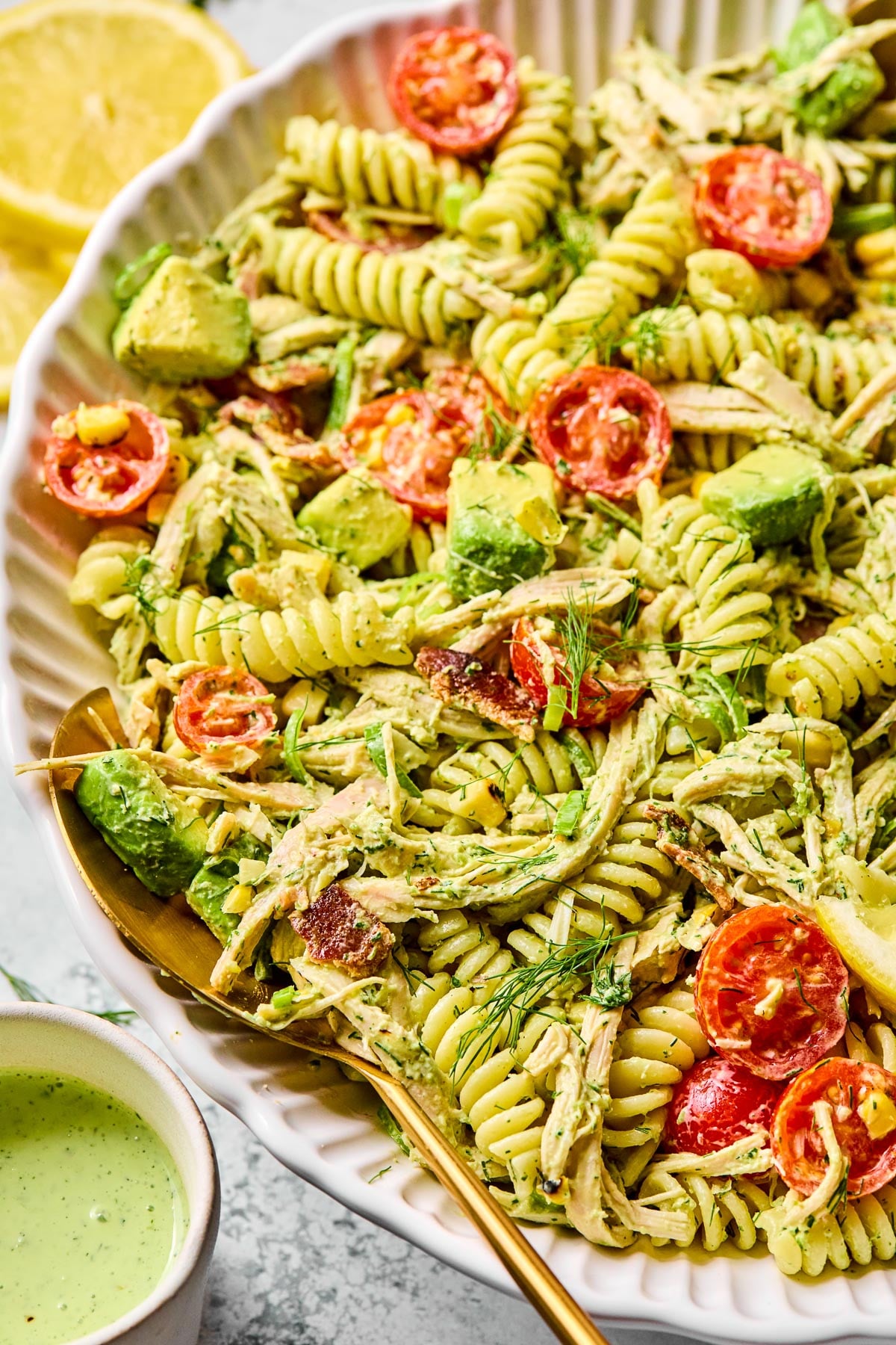 Chicken bacon ranch pasta salad in a large white bowl. There is a spoon in the bowl and it pasta salad is garnished with dill. Around the bowl is a small cup of jalapeño ranch and lemon slices.