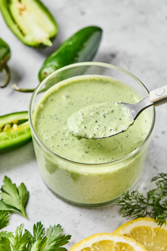 Jalapeño ranch in a glass cup with a spoonful being held over it. Around the cup are fresh dill, some slices of lemon, fresh parsley, and some jalapeños.